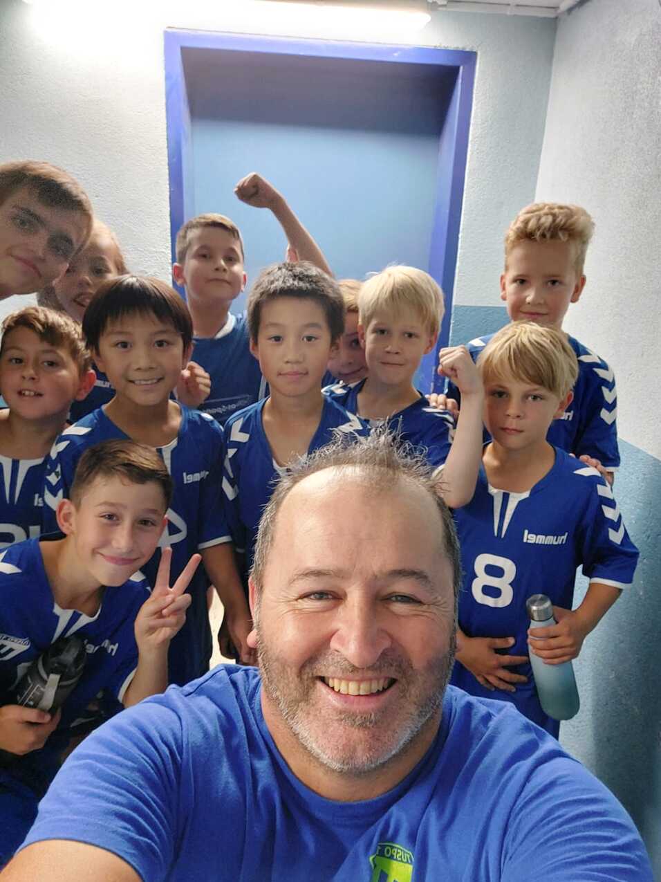Gruppenselfie von einem Trainer und zehn Jungen in blauen Trikots in einer Umkleide. Freude und Zusammenhalt sind spürbar.