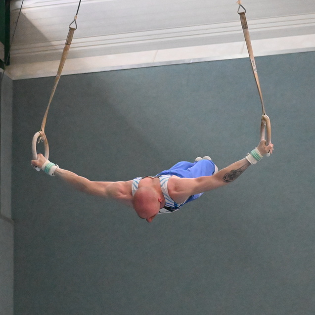 Turner beim Schwingen an Ringen, in blauer Sportkleidung, mit gestrecktem K&ouml;rper und konzentrierter K&ouml;rperhaltung.