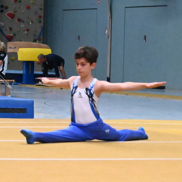 Junge ausf&uuml;hren einer Spagat&uuml;bung auf einem Turnboden in einer Sporthalle, Kulisse mit Kletterwand im Hintergrund.