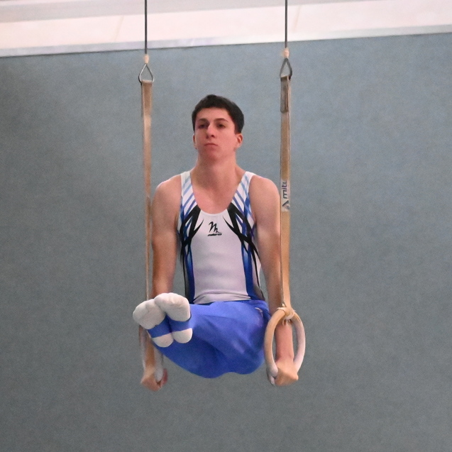Junger Turner sitzt in einer stabilen Position an Ringen in einer Turnhalle mit grauem Hintergrund.