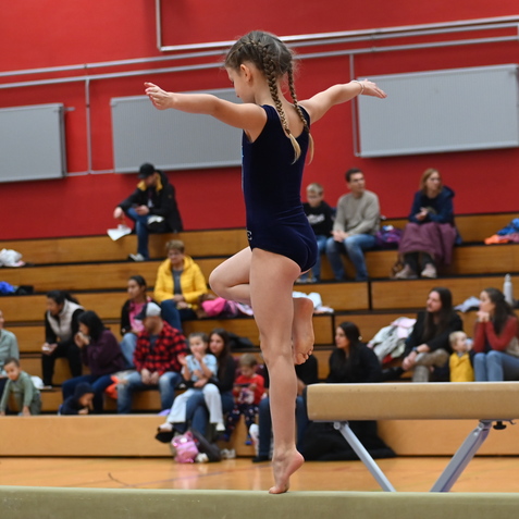 M&auml;dchen in Turnanzug balanciert auf einem Schwebebalken, Zuschauer im Hintergrund auf Trib&uuml;nen.