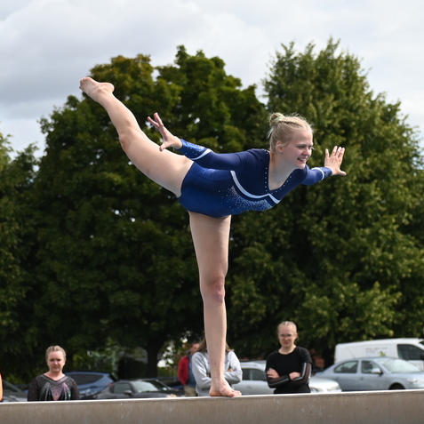 Turnerin in blauer Sportskleidung balanciert auf einem Schwebebalken, umgeben von Zuschauern und B&auml;umen.