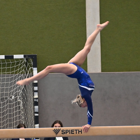 Turnerin in blauer Gymnastikanzug macht Handstand auf dem Schwebebalken, im Hintergrund Zuschauer und ein Tor.