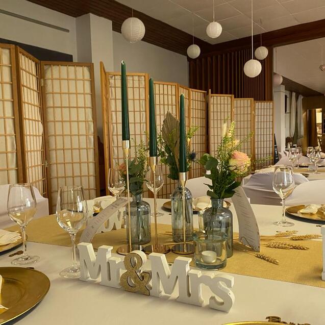 Elegant gedeckter Tisch mit „Mr & Mrs“-Dekoration, Blumenarrangements und Gläsern, umgeben von einem Raumteiler.