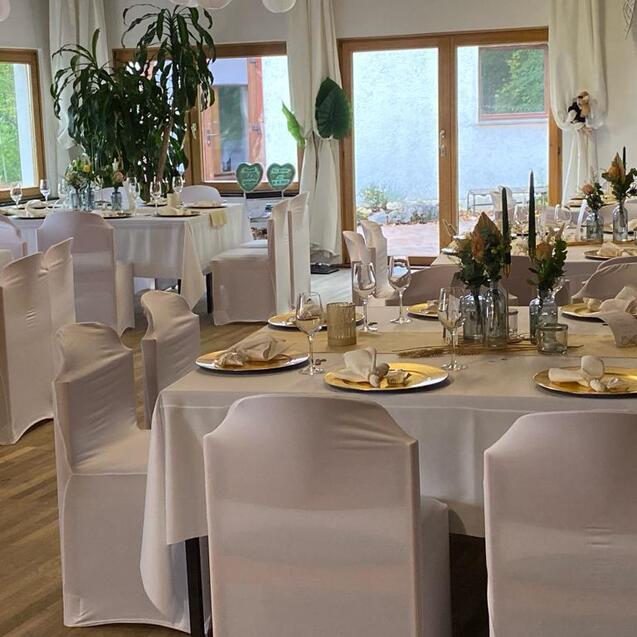 Festlich dekorierter Veranstaltungsraum mit weißen Tischen, Stühlen und Blumenarrangements. Große Fenster sorgen für Tageslicht.