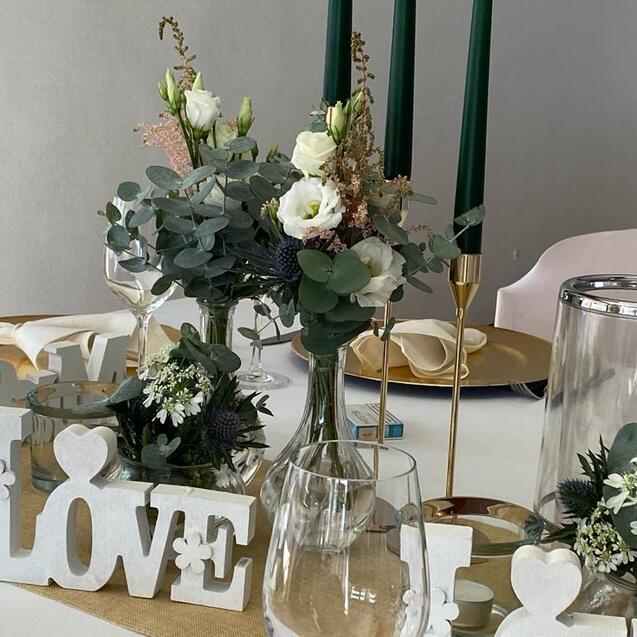Tischdeko mit grünen Kerzen, Blumenarrangements, Gläsern und einer „LOVE“-Dekoration auf einem eleganten Tisch.