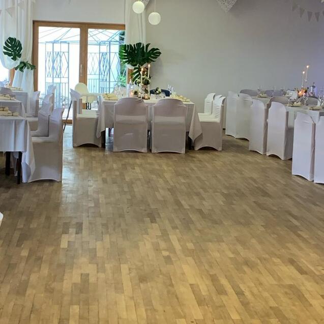 Festlich dekorierter Veranstaltungsraum mit weißen Tischen, Stühlen und Pflanzen, Holzfußboden und viel natürlichem Licht.
