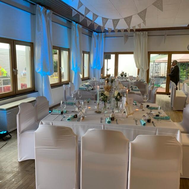 Festlich dekorierter Saal mit gedeckten Tischen, weißen Stuhlhussen und Lichtern, ideal für Veranstaltungen oder Feiern.