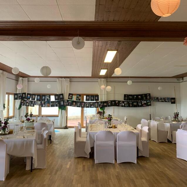 Festlich dekorierter Saal mit gedeckten Tischen, weißen Stühlen und Bildern an der Wand, ideal für Veranstaltungen.