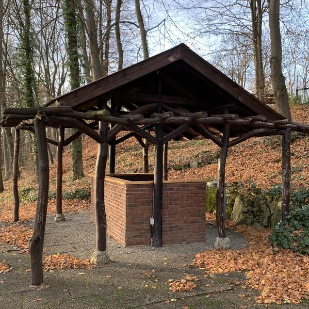 Pavillon aus Holz mit Ziegelsäule in herbstlicher Umgebung mit buntem Laub am Boden.