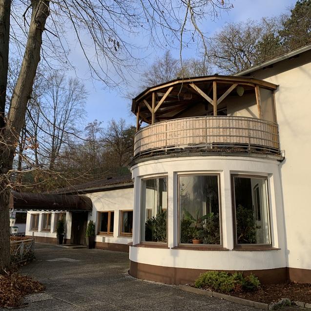 Ansicht eines Hauses mit verglaster Rundbank und Balkon, umgeben von Bäumen und einem gepflasterten Weg.