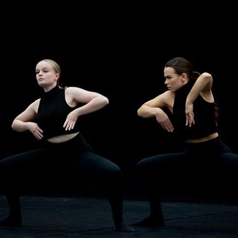 Zwei Tänzerinnen in schwarzer Kleidung führen synchronisierte Bewegungen auf einer dunklen Bühne aus.