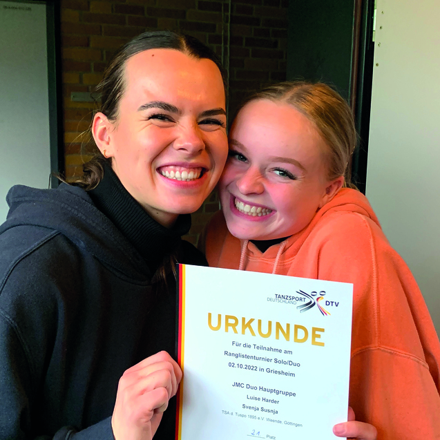 Zwei junge Frauen l&auml;cheln freudig, w&auml;hrend sie eine Urkunde f&uuml;r einen Tanzwettbewerb halten.