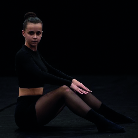 T&auml;nzerin sitzt auf dem Boden in schwarzem Outfit vor dunklem Hintergrund, mit ruhigem Gesichtsausdruck.