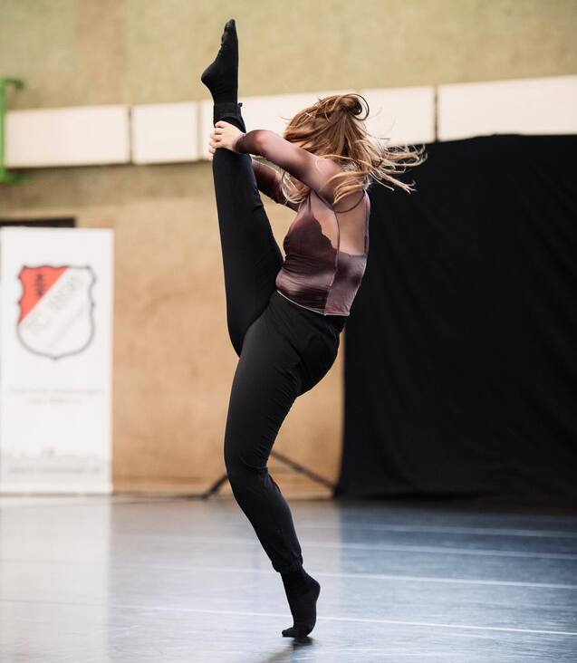 T&auml;nzerin in schwarzer Leggings und top mit ausgestrecktem Bein in der Luft, Hintergrund mit unscharfem Banner.