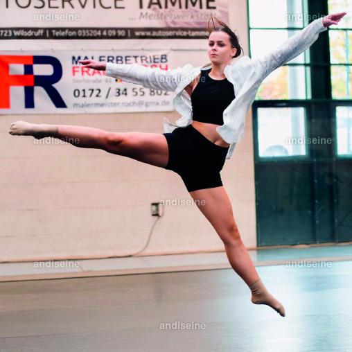 T&auml;nzerin f&uuml;hrt einen kraftvollen Sprung in einer Trainingshalle aus, mit ausgestreckten Armen und einer modernen Sportkleidung.