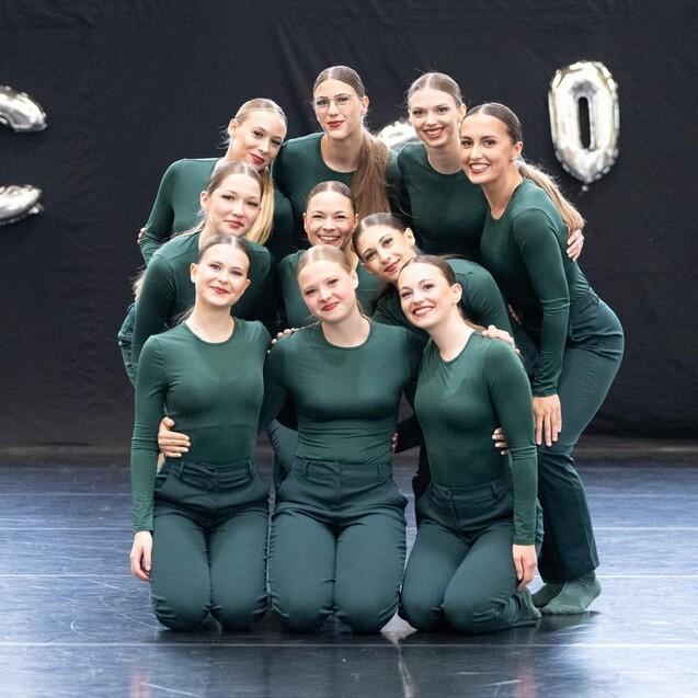 Gruppierung von neun T&auml;nzerinnen in gr&uuml;ner Kleidung, die l&auml;chelnd auf einer B&uuml;hne posieren, im Hintergrund silberne Luftballons.