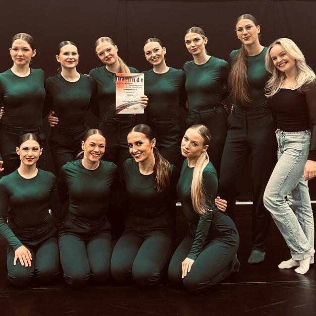 Gruppe von zehn t&auml;nzerischen Darstellern in dunkelgr&uuml;ner Kleidung posierend mit einer Auszeichnung in der Mitte.