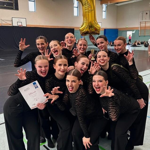 Gruppe von T&auml;nzerinnen in schwarzen Outfits posiert um einen Pokal und eine goldene &bdquo;1&ldquo;-Ballon, l&auml;chelnd und feiernd.