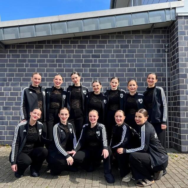 Gruppe von zw&ouml;lf T&auml;nzerinnen in schwarz-wei&szlig;en Outfits, posierend vor einer grauen Wand unter blauem Himmel.