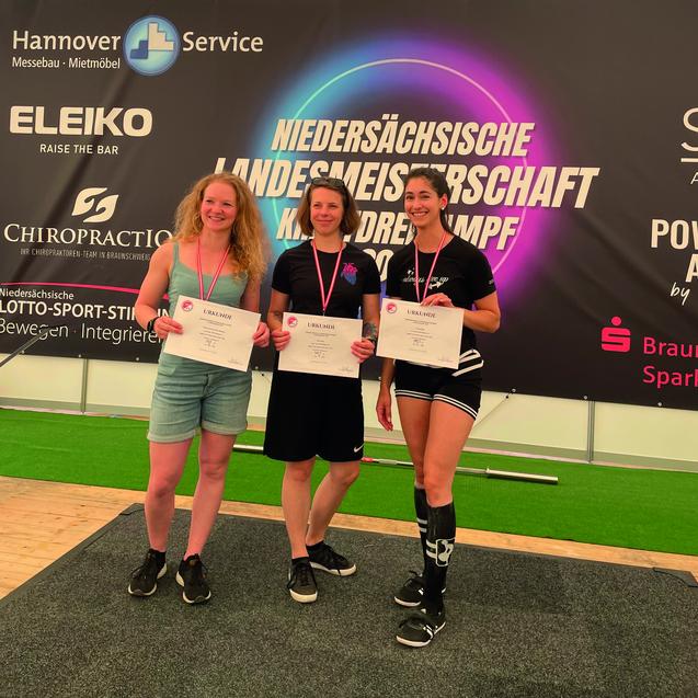 Drei Frauen mit Medaillen stehen l&auml;chelnd vor einem Banner der Nieders&auml;chsischen Landesmeisterschaft Kraftdreikampf.