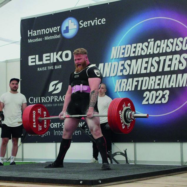 Starker Gewichtheber hebt eine Langhantel mit roten Gewichten, begleitet von zwei Zuschauern bei einem Wettkampf.