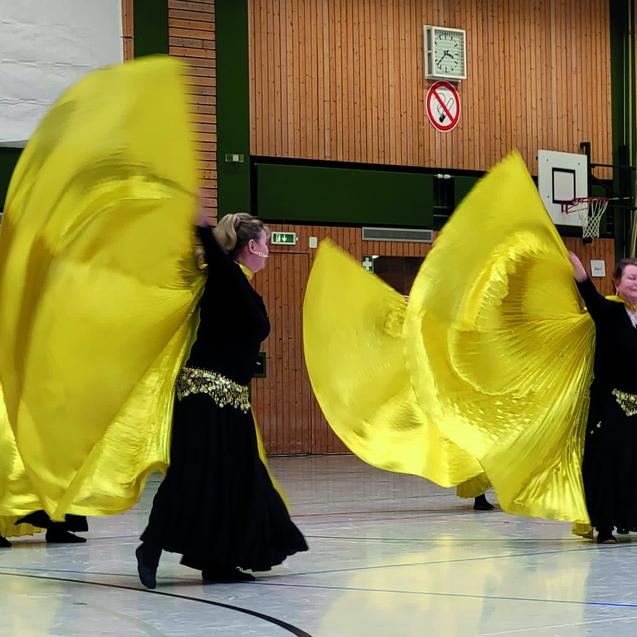 Tanzende Frauen in schwarzen Kost&uuml;men schwenken gro&szlig;e gelbe Schleier in einer Sporthalle.