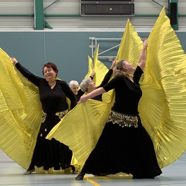 Tanzende Frauen in schwarzen Kleidern schwingen gro&szlig;e, gelbe Faltstoffe in einer Sporthalle.