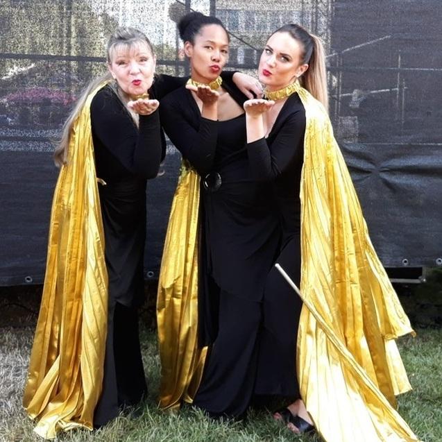 Drei Frauen in schwarzen Outfits mit goldenen Capes posieren l&auml;chelnd und k&uuml;ssen die Kamera.