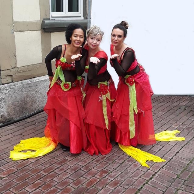 Drei Frauen in roten Kost&uuml;men mit bl&uuml;henden Verzierungen posieren fr&ouml;hlich auf einem Pflasterweg.