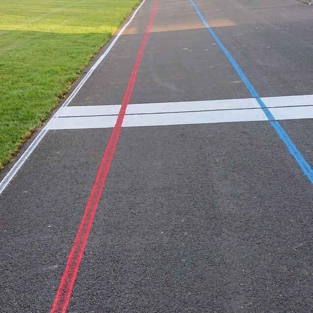 Kurve eines Radrennbahn mit roten und blauen Markierungen, die zur Ziellinie f&uuml;hren, umgeben von gr&uuml;nem Rasen.