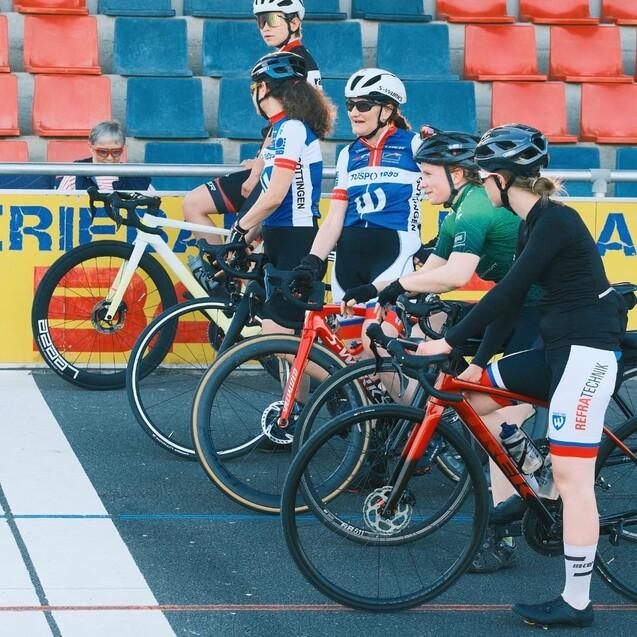Sechs Radfahrerinnen in sportlicher Kleidung stehen an der Startlinie einer Radbahn, bereit zum Wettkampf.