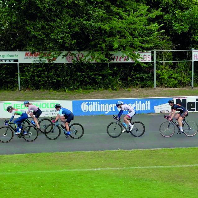 Gruppe von Radfahrern in Rennbekleidung f&auml;hrt auf einer velodromartigen Strecke mit Werbebannern im Hintergrund.