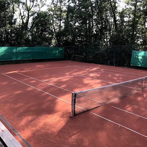 Tennisplatz mit rotem Sandbelag, Netz in der Mitte und umgeben von B&auml;umen und gr&uuml;nen Sichtschutzw&auml;nden.