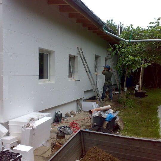 Bauarbeiter installiert D&auml;mmplatten an einer wei&szlig; gestrichenen Hauswand im Garten, umgeben von Werkzeug und Materialien.