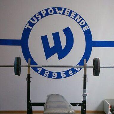 Langhantel auf einem Trainingsstand neben einer Wand mit dem Logo von TuS Powewende.