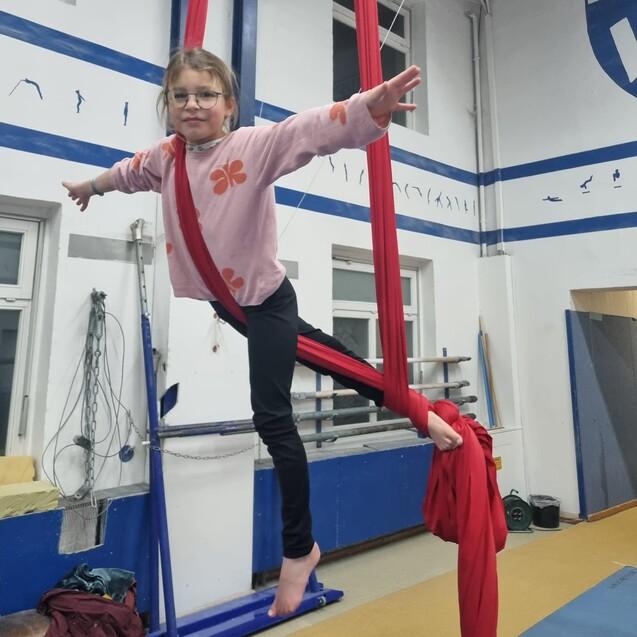 M&auml;dchen in rosa Pullover schwingt an roten T&uuml;chern in einer Sporthalle, mit ausgebreiteten Armen und l&auml;chelndem Gesicht.