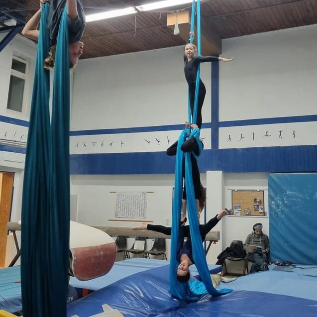 Drei Personen turnen an blauen Luftakrobatik-T&uuml;chern in einer Turnhalle &uuml;ber einer Landematte.