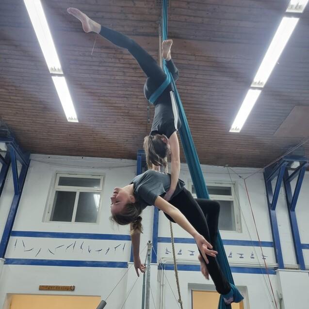 Zwei Akrobatinnen h&auml;ngen an blauen T&uuml;chern und vollziehen eine koordinierte &Uuml;bung in einer Sporthalle.