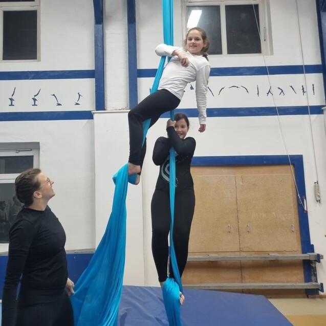Drei Personen mit Lufthandstand an blauen Tuchschlingen in einer Turnhalle, eine Person sitzt auf einem Tuch.