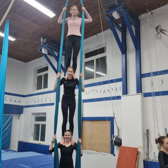 Drei Frauen stehen auf &uuml;bereinander gestapelten T&uuml;chern in einer Turnhalle, gesichert von blauen T&uuml;chern und Matten.