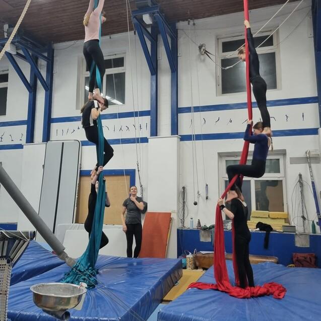 Sechs Artistinnen &uuml;ben an bunten T&uuml;chern in einer Zirkus-Trainingseinrichtung mit Matten im Hintergrund.