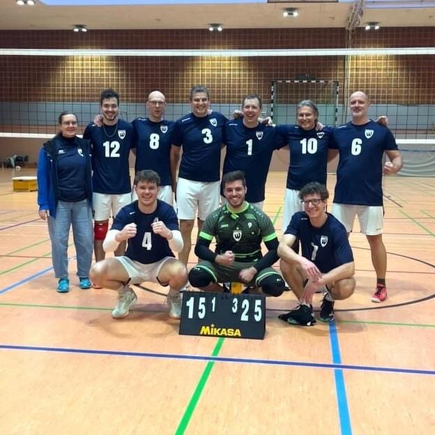 Gruppe von Volleyballspielern in Vereinsanz&uuml;gen posiert auf dem Feld vor einer Anzeigetafel mit dem Ergebnis 15:1 und 32:5.