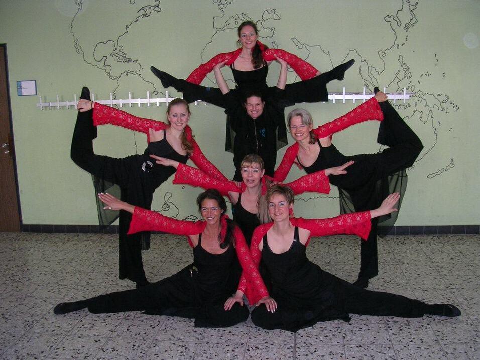 Gruppenszene mit sieben T&auml;nzerinnen in schwarzer Kleidung und roten &Auml;rmeln, die akrobatische Posen auf einem Boden einnehmen.