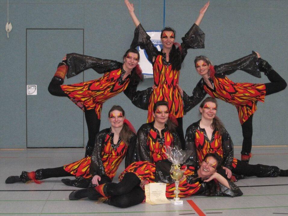 Gruppe von T&auml;nzerinnen in auff&auml;lligen, feuerfarbenen Kost&uuml;men, posierend mit Pokal und Urkunde auf einer Sporthalle.