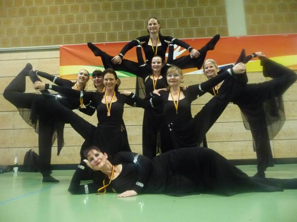 Acht T&auml;nzerinnen in schwarzen Kost&uuml;men posieren kreativ auf einer gr&uuml;nen Fl&auml;che. Medaillen h&auml;ngen um ihren Hals.