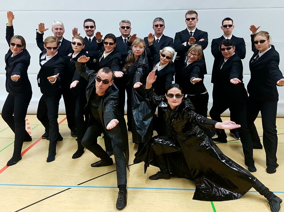 Gruppe von 18 Personen in Anz&uuml;gen mit Sonnenbrillen posiert dynamisch in einem Sporthallenraum.