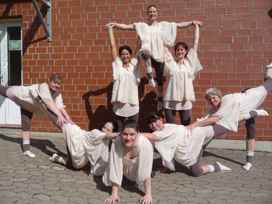 Acht Frauen in eleganten, hellen Outfits posieren kreativ vor einer Backsteinwand.