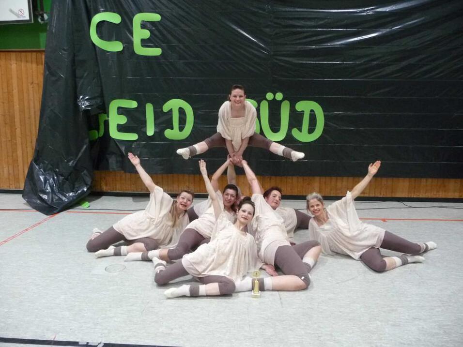 Gruppe von sechs T&auml;nzerinnen posiert kreativ auf dem Boden vor einer Wand mit dem Schriftzug &bdquo;CE leid S&uuml;d&ldquo;.