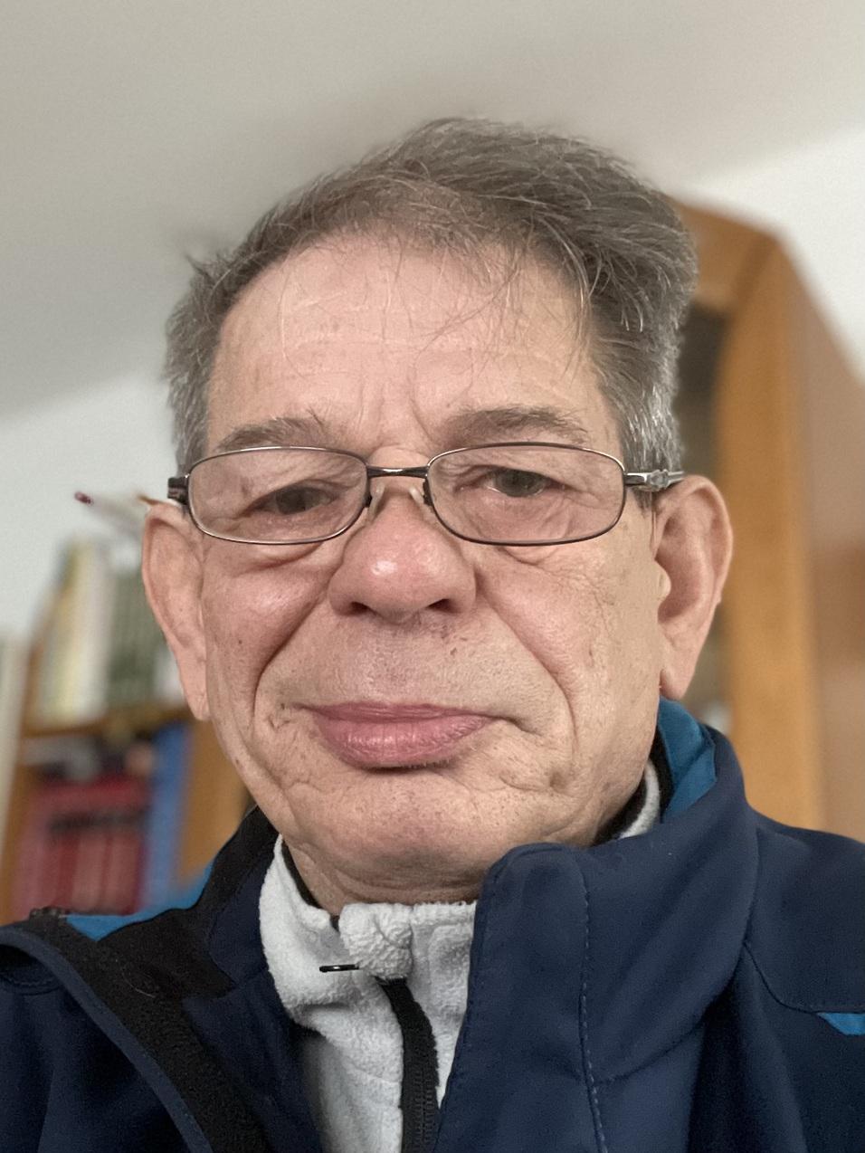 Porträt eines älteren Mannes mit Brille, lächelnd, in einer dunklen Jacke vor einem Bücherregal.